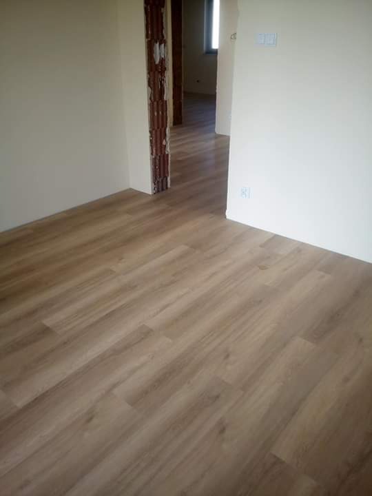 Jasnobrązowe panele podłogowe ułożone w nowym pomieszczeniu z widocznymi fragmentami surowej cegły w otworze drzwiowym i świeżo pomalowanymi ścianami w odcieniach beżu.