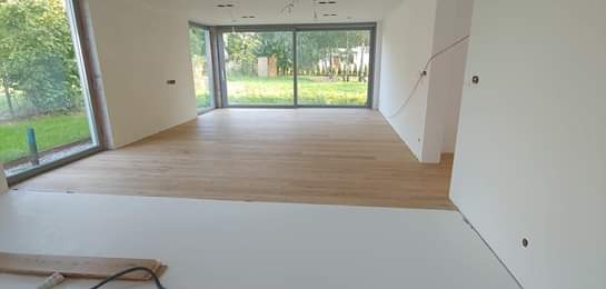 Jasny parkiet ułożony we wnętrzu z dużymi oknami, widoczne przygotowania do dalszych prac wykończeniowych, w tle zielony ogród.