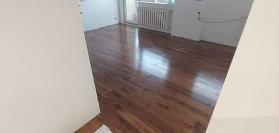 Błyszcząca, świeżo położona drewniana podłoga w pokoju z białymi ścianami i grzejnikiem pod oknem. Widoczne niedoskonałości przy ścianach, wskazujące na trwający remont.