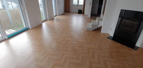 Jasny parkiet ułożony we wzór jodełki w przestronnym salonie w Katowicach, z widocznym kominkiem i schodami w tle.