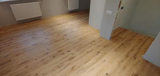 Jasne panele podłogowe o strukturze drewna, ułożone w nowym mieszkaniu, widoczny grzejnik i fragment ściany w szarym odcieniu.