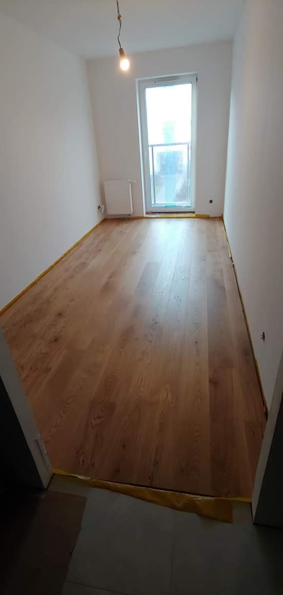 Wykończone wnętrze z drewnianą podłogą, zabezpieczoną żółtą taśmą malarską wzdłuż ścian i progu, surowa żarówka zwisająca z sufitu.