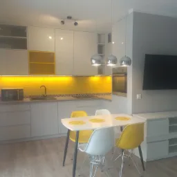 Nowoczesna, jasna kuchnia z żółtymi akcentami: podświetlane szafki, mozaika nad blatem i krzesła przy białym stole jadalnym, oświetlona trzema chromowanymi lampami wiszącymi.