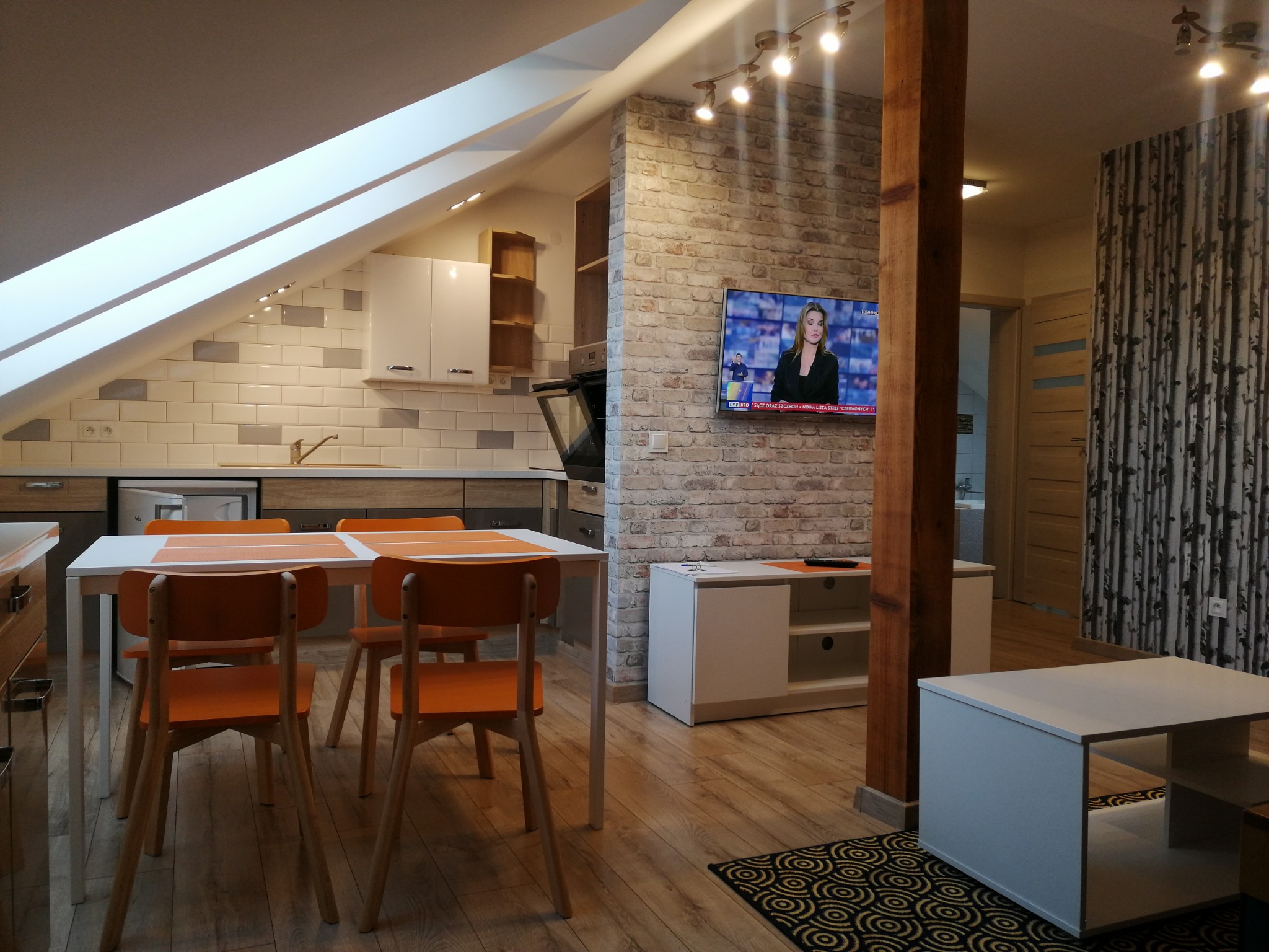 Wnętrze mieszkania na poddaszu z jadalnią, kuchnią i salonem, ceglana ściana z telewizorem, drewniana kolumna i podłoga, pomarańczowe krzesła przy białym stole.