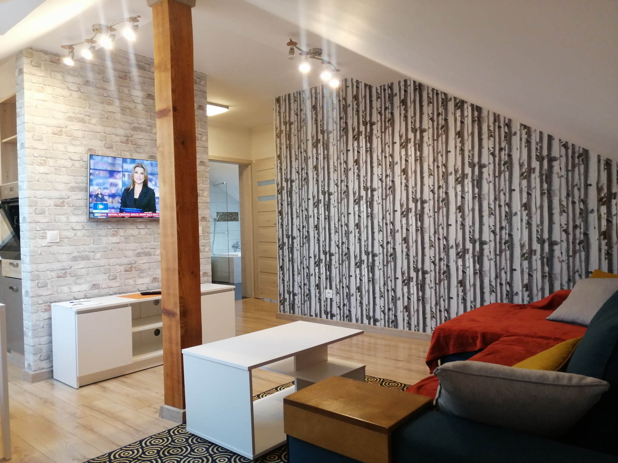 Salon z ceglaną ścianą, telewizorem, białą szafką RTV, drewnianym słupem, biało-brązowym stolikiem kawowym i ścianą z tapetą w motywie brzozowego lasu.