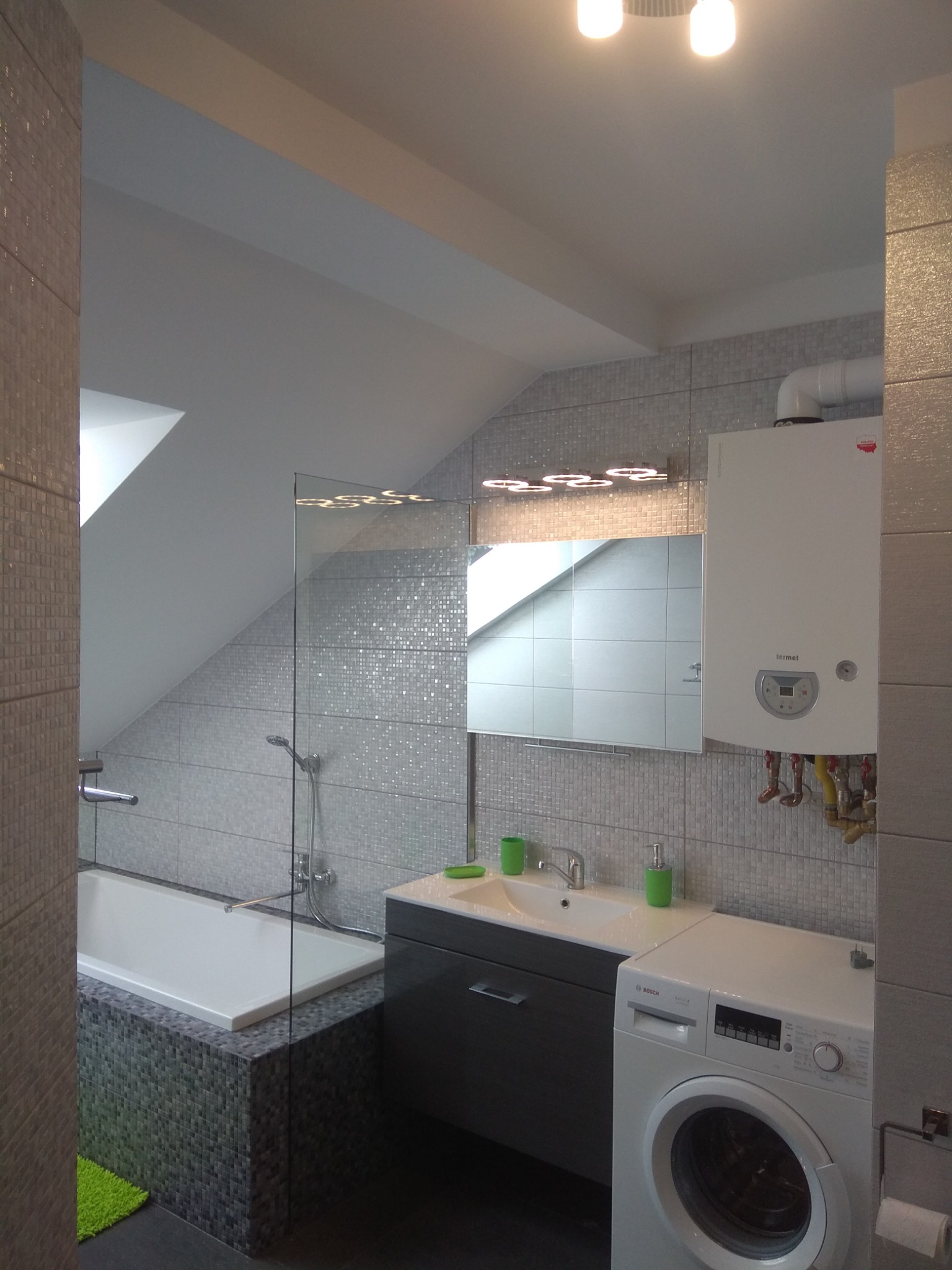 Nowoczesna łazienka na poddaszu ze skośnym sufitem, wykończona mozaiką w odcieniach szarości, z białą wanną obudowaną mozaiką, umywalką z szafką, pralką Bosch i wiszącym piecem gazowym Termet.