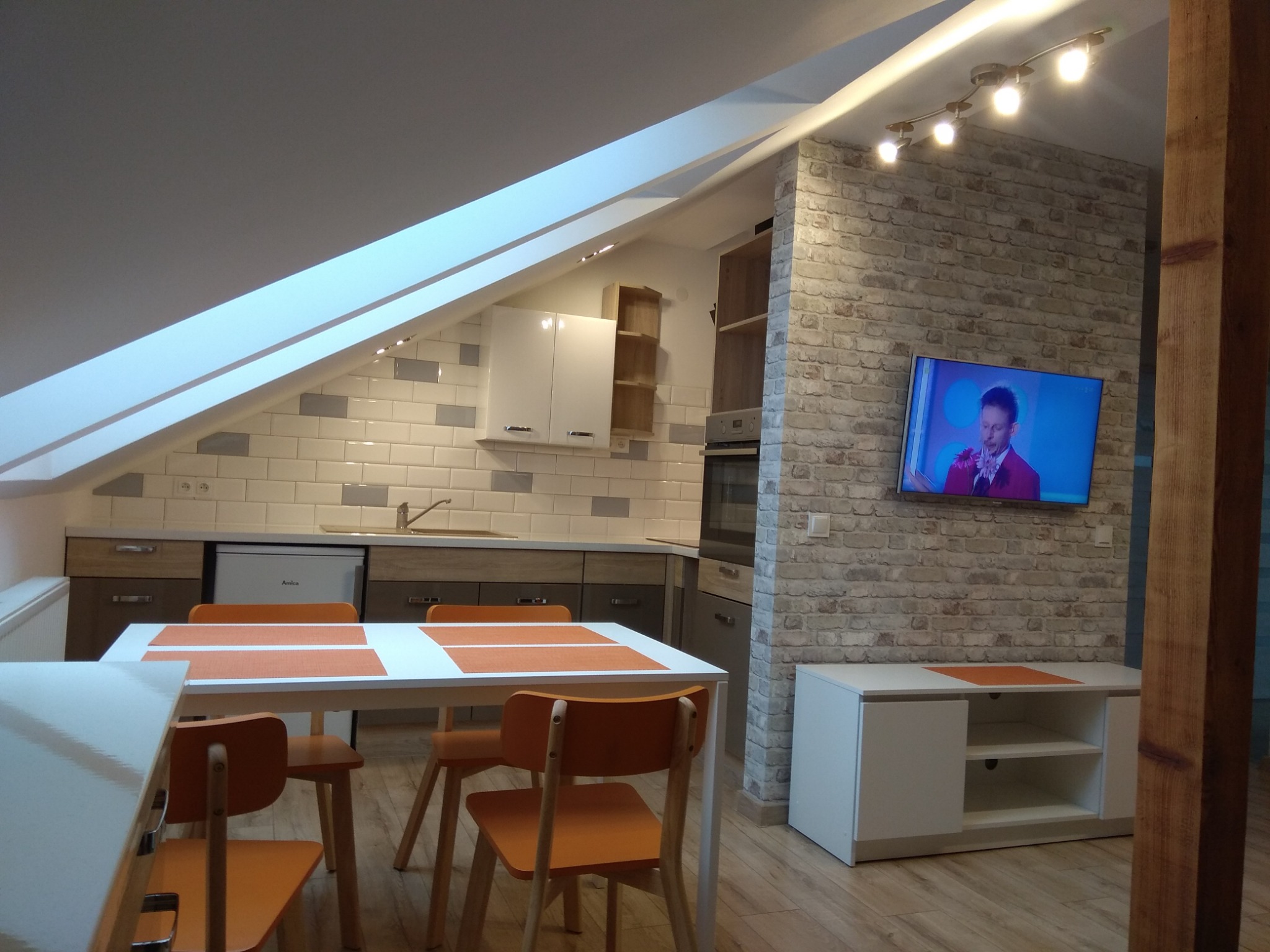 Jasna kuchnia w mieszkaniu na poddaszu z białymi meblami, drewnianymi akcentami i ceglaną ścianą z telewizorem, widok na stół z pomarańczowymi krzesłami.