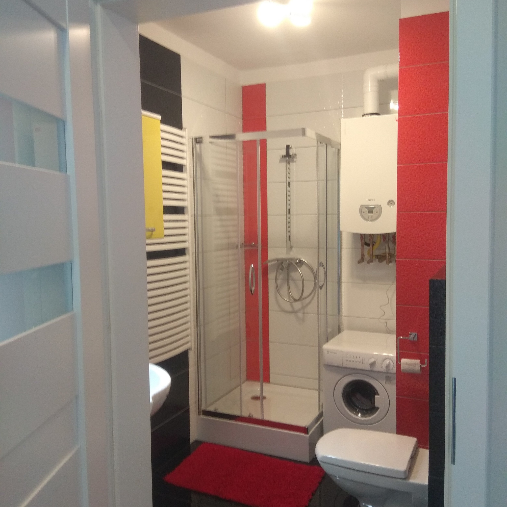 Wnętrze łazienki po remoncie: kabina prysznicowa z czerwoną listwą, grzejnik drabinkowy z żółtym ręcznikiem, pralka obok toalety, czerwony dywanik na czarnych płytkach podłogowych.