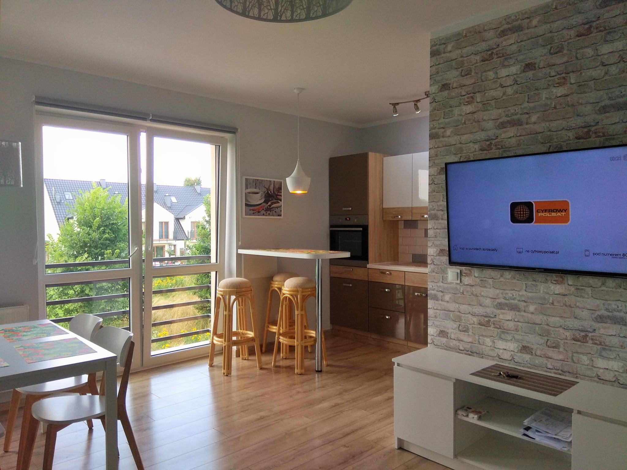 Jasne wnętrze mieszkania z parkietem, ceglaną ścianą z telewizorem, stołem z krzesłami barowymi i widokiem na zieleń za oknem balkonowym.