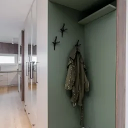 Nowoczesny przedpokój z szafą wnękową, lustrzanymi drzwiami i siedziskiem w pepitkę, zielona wnęka z wieszakami w kształcie gałęzi i wiszącą kurtką.