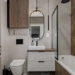 Nowoczesna łazienka z płytkami imitującymi marmur, owalnym lustrem z podświetleniem LED, wiszącą szafką z drewnianymi frontami oraz białą umywalką nablatową i baterią w kolorze czarnym.