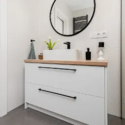 Nowoczesna łazienka z białą szafką pod umywalkę z drewnianym blatem, okrągłym lustrem w czarnej ramie i czarną armaturą, uzupełniona o minimalistyczne dodatki i oświetlenie.