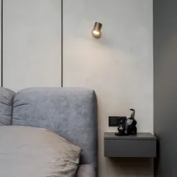 Szare tapicerowane wezgłowie łóżka z poduszką, minimalistyczna szafka nocna z czarną dekoracyjną figurką i gniazdkiem elektrycznym, oświetlenie punktowe na ścianie w odcieniach szarości z czarnymi...