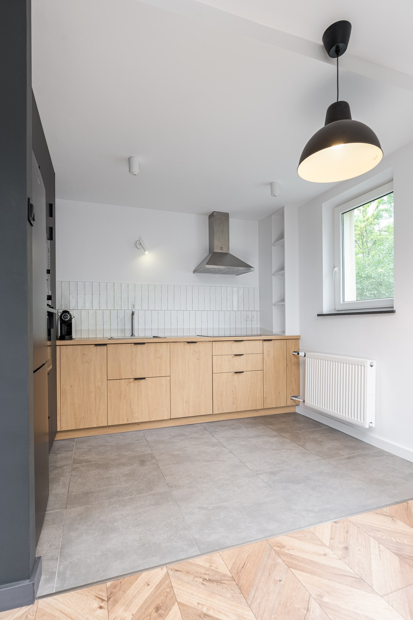 Jasna kuchnia z drewnianymi frontami szafek, białymi płytkami nad blatem i szarymi płytkami podłogowymi, widoczny okap, okno i czarna lampa sufitowa.