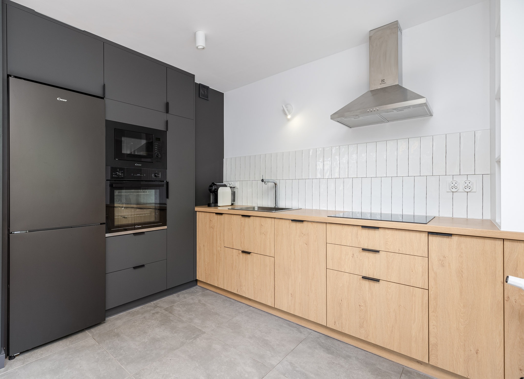 Nowoczesna kuchnia: grafitowe szafki z zabudowanym sprzętem AGD (lodówka, piekarnik, mikrofalówka) kontrastują z jasnymi drewnianymi frontami szafek dolnych i białymi płytkami na ścianie.