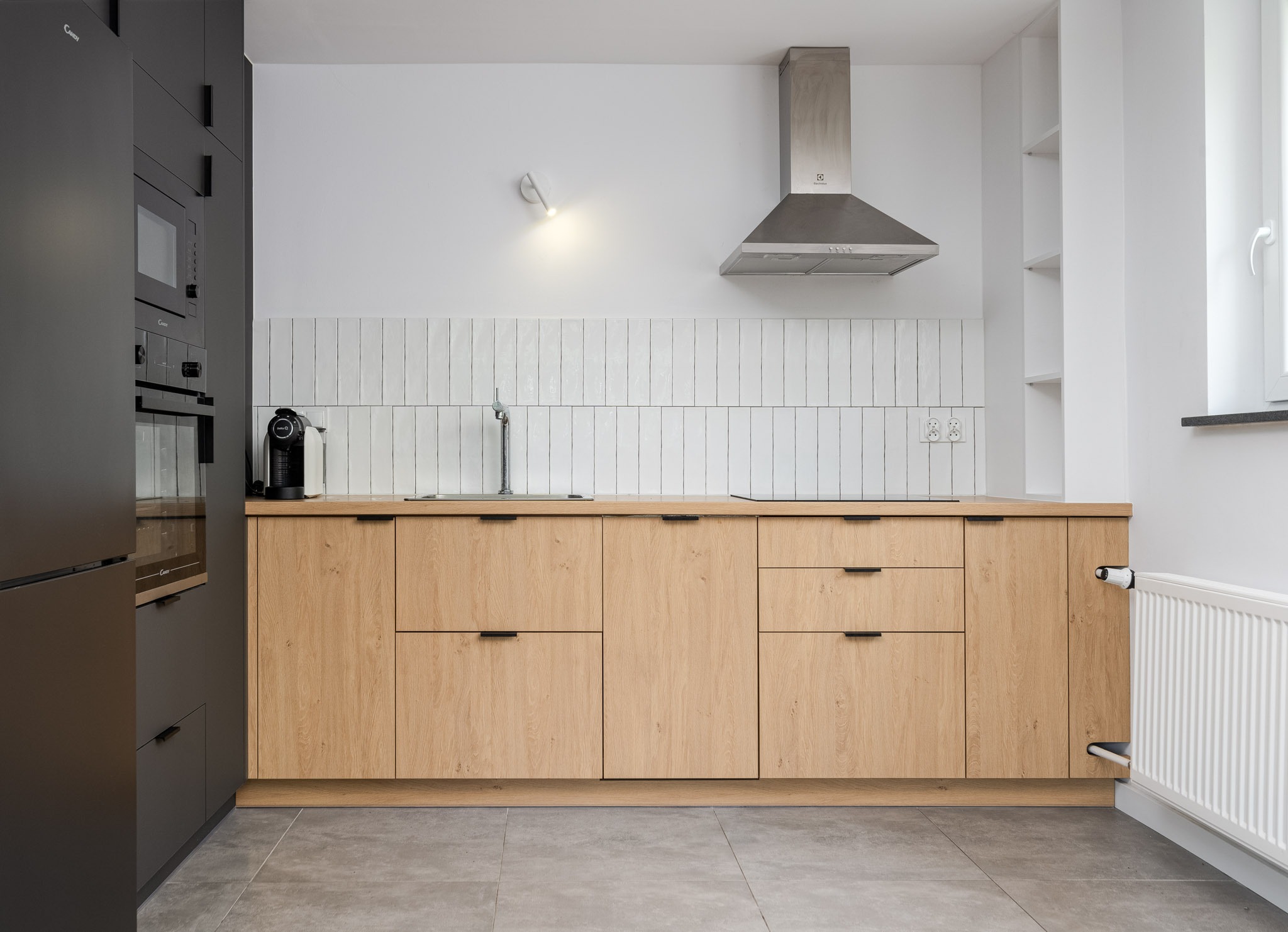 Nowoczesna kuchnia z drewnianymi frontami szafek, czarnymi uchwytami i białymi, pionowo ułożonymi płytkami na ścianie. Widoczny fragment zabudowy z piekarnikiem i mikrofalówką w czarnym kolorze...