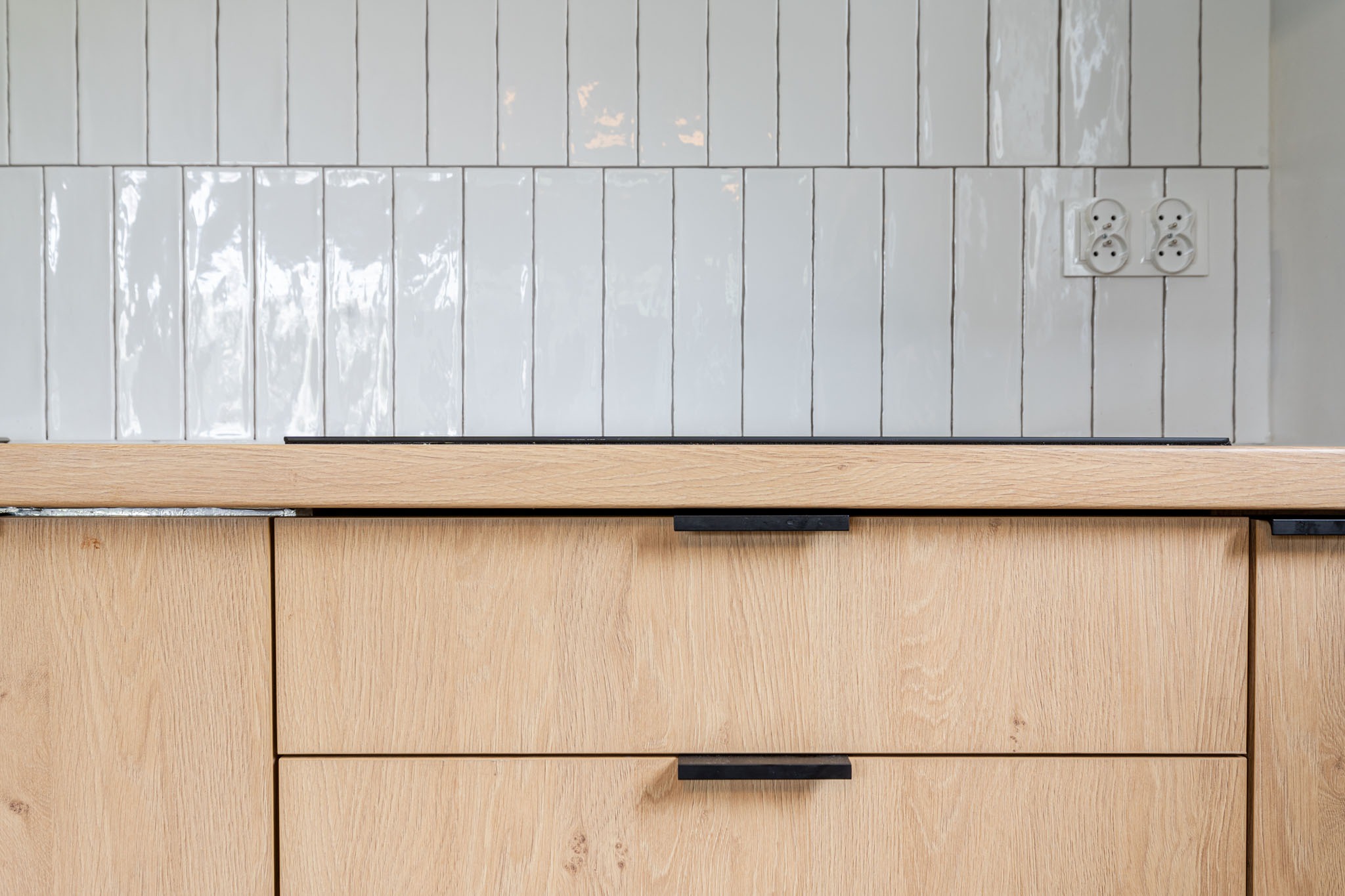 Fragment zabudowy kuchennej z drewnianym blatem i frontami szuflad w odcieniach jasnego drewna, czarne, minimalistyczne uchwyty, tło z białych, błyszczących płytek ułożonych pionowo, widoczne...