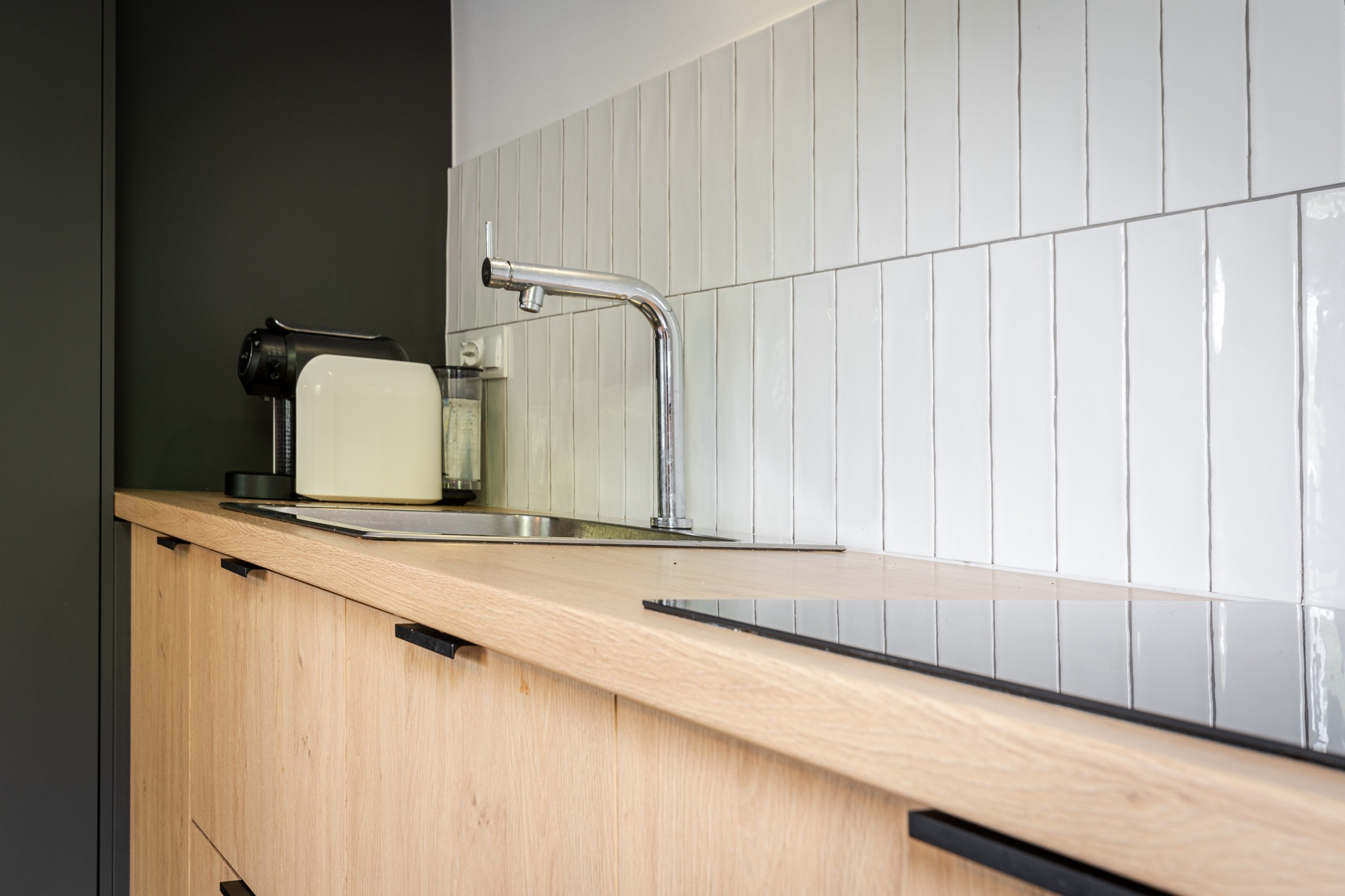 Jasny drewniany blat kuchenny z czarnymi uchwytami szafek, chromowaną baterią i białymi pionowymi płytkami na ścianie, uzupełniony czarnym ekspresem do kawy.