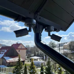 Dwie czarne kamery przemysłowe zamontowane pod drewnianym zadaszeniem, widok na zasypany śniegiem krajobraz wiejski z budynkami w tle, błękitne niebo z chmurami.