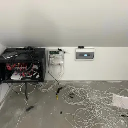 Niedokończona instalacja sieci komputerowej w pomieszczeniu ze skośnym sufitem, widoczna szafa rack z okablowaniem, rozłożone kable na podłodze, skrzynka rozdzielcza na ścianie i narzędzia.