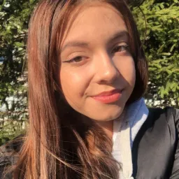 Portret uśmiechniętej młodej kobiety z brązowymi włosami na tle zielonych gałęzi drzewa.