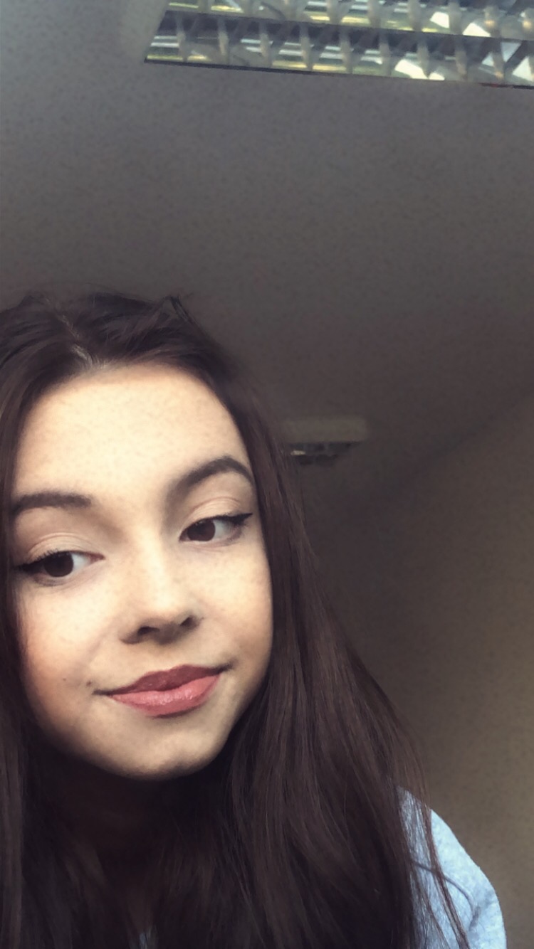 Portret młodej kobiety z ciemnymi włosami i delikatnym makijażem, patrzącej w lewo z lekkim uśmiechem, z widocznym oświetleniem sufitowym w tle.