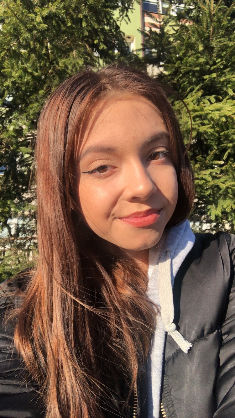 Portret uśmiechniętej młodej kobiety z brązowymi włosami na tle zielonych gałęzi drzewa.