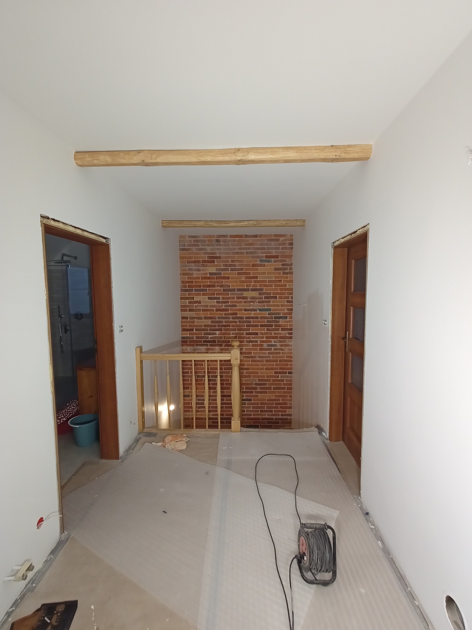 Wnętrze w trakcie remontu: widoczne surowe drewniane belki pod sufitem, fragment ściany wyłożony cegłą, zamontowane ościeżnice drzwiowe, podłoga zabezpieczona folią, zwinięty przedłużacz elektryczny.