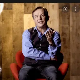 Mężczyzna w niebieskiej koszuli i kamizelce siedzi na czerwonym krześle, gestykulując dłonią z zegarkiem na tle drewnianej ściany z geometrycznymi wzorami.