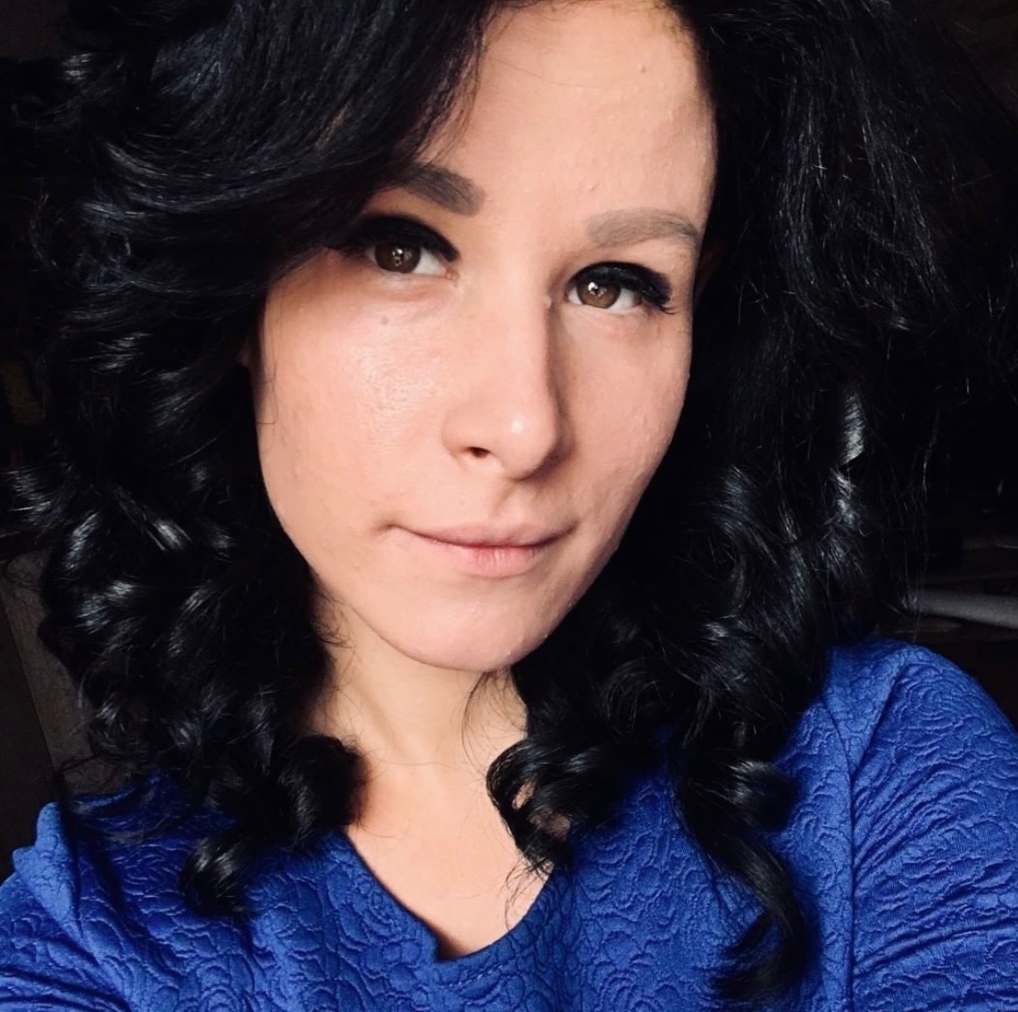 Portret kobiety w niebieskiej bluzce z koronkowym wzorem i z czarnymi, kręconymi włosami, z delikatnym uśmiechem.