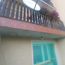 Kątowe ujęcie balkonu z brązową, drewnianą balustradą, na kt&oacute;rej zamontowana jest antena satelitarna i donica z fioletowymi kwiatami; widoczne zabezpieczone folią okno na elewacji budynku z fakturą.