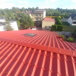 Czerwony dach z blachy trapezowej z oknem dachowym, widok z góry na tle zabudowy jednorodzinnej i zieleni.