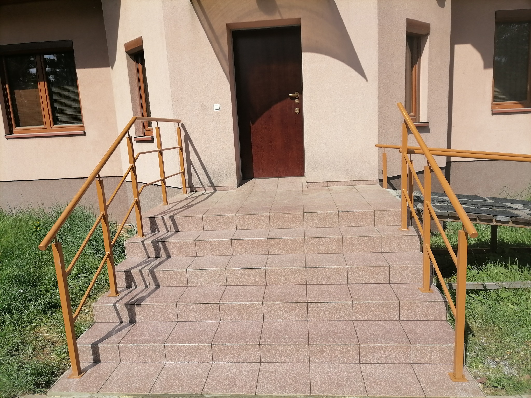 Zewnętrzne schody wejściowe do budynku, wyłożone płytkami w odcieniach beżu i brązu, z metalową, pomalowaną na złoto-brązowo barierką. Widoczna drewniana stolarka okienna i drzwi wejściowe...