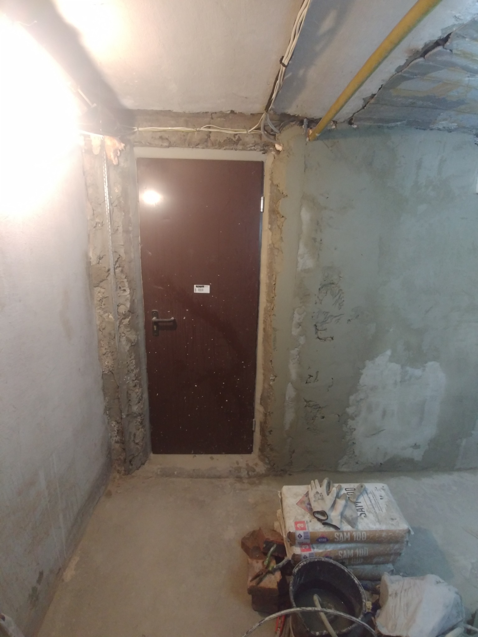 Przygotowanie do prac wykończeniowych w pomieszczeniu: worki z zaprawą SAM 100, wiadro, rękawice robocze i narzędzia leżą na podłodze, ściany surowe po tynkowaniu, widoczna instalacja elektryczna...