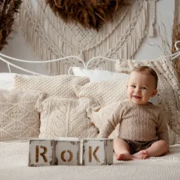 Uśmiechnięty roczny chłopiec siedzi na łóżku w beżowym swetrze i spodenkach, z drewnianymi klockami z napisem 'ROK' przed sobą, dekoracje z suszonych traw i makramy w tle.