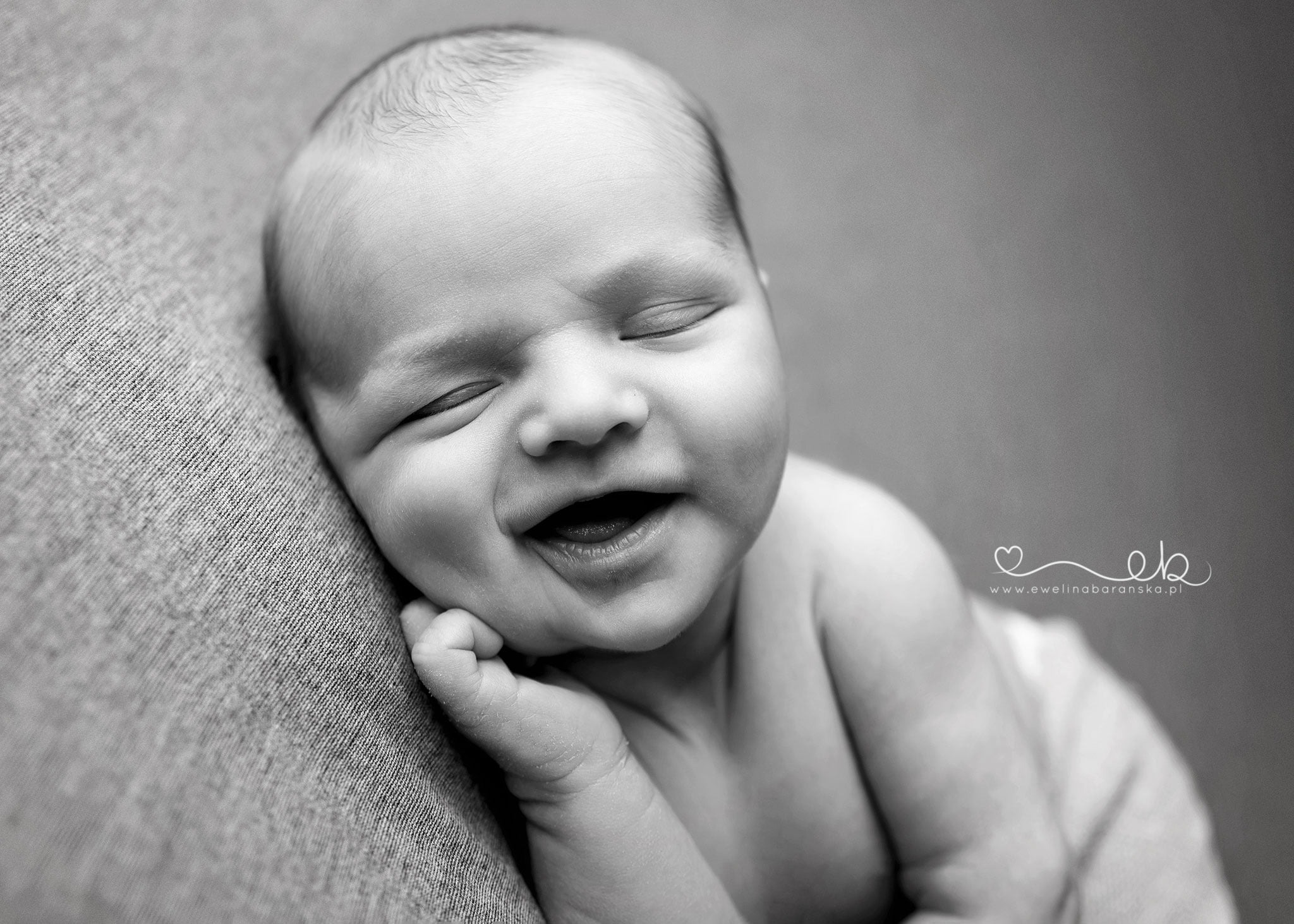 Czarno-białe zbliżenie śpiącego noworodka z uśmiechem na twarzy, opierającego policzek na dłoni, na teksturowanym tle, z widocznym logo fotografa.