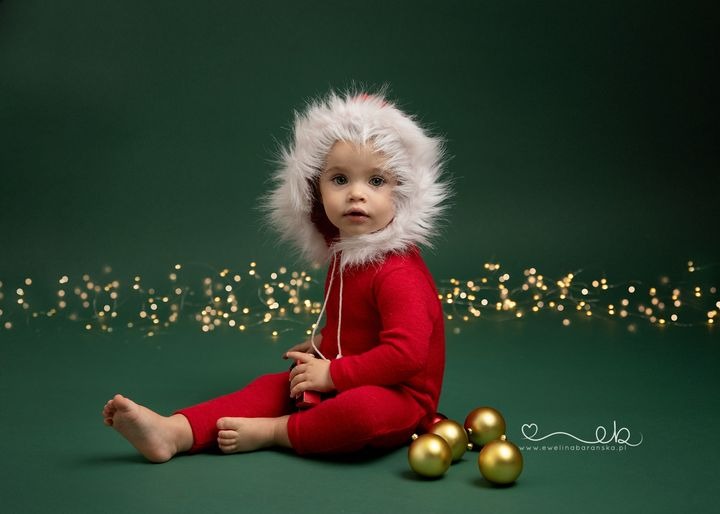Radosny maluch w czerwonym kombinezonie z futrzanym kapturem, siedzący na zielonym tle z rozproszonymi światełkami i złotymi bombkami, uchwycony w studiu fotograficznym z logo fotografa w prawym...