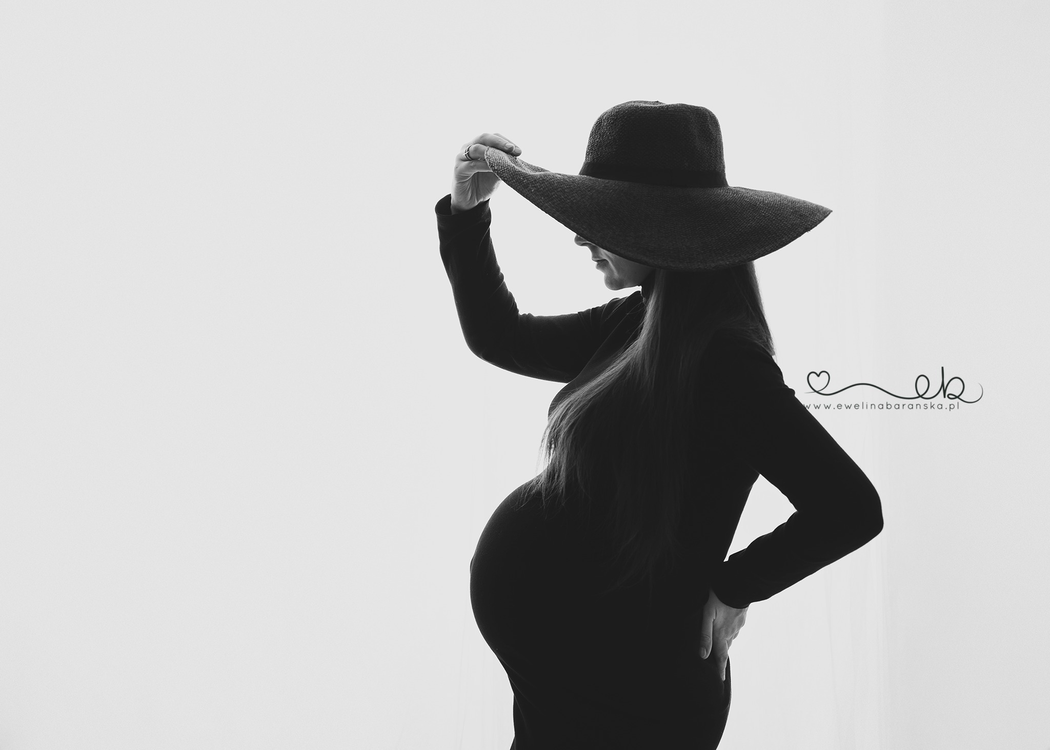 Czarno-białe zdjęcie sylwetki kobiety w ciąży, pozującej z kapeluszem z szerokim rondem, dłoń uniesiona do ronda, strój z długim rękawem, widoczny profil i zaokrąglony brzuch na jasnym tle.