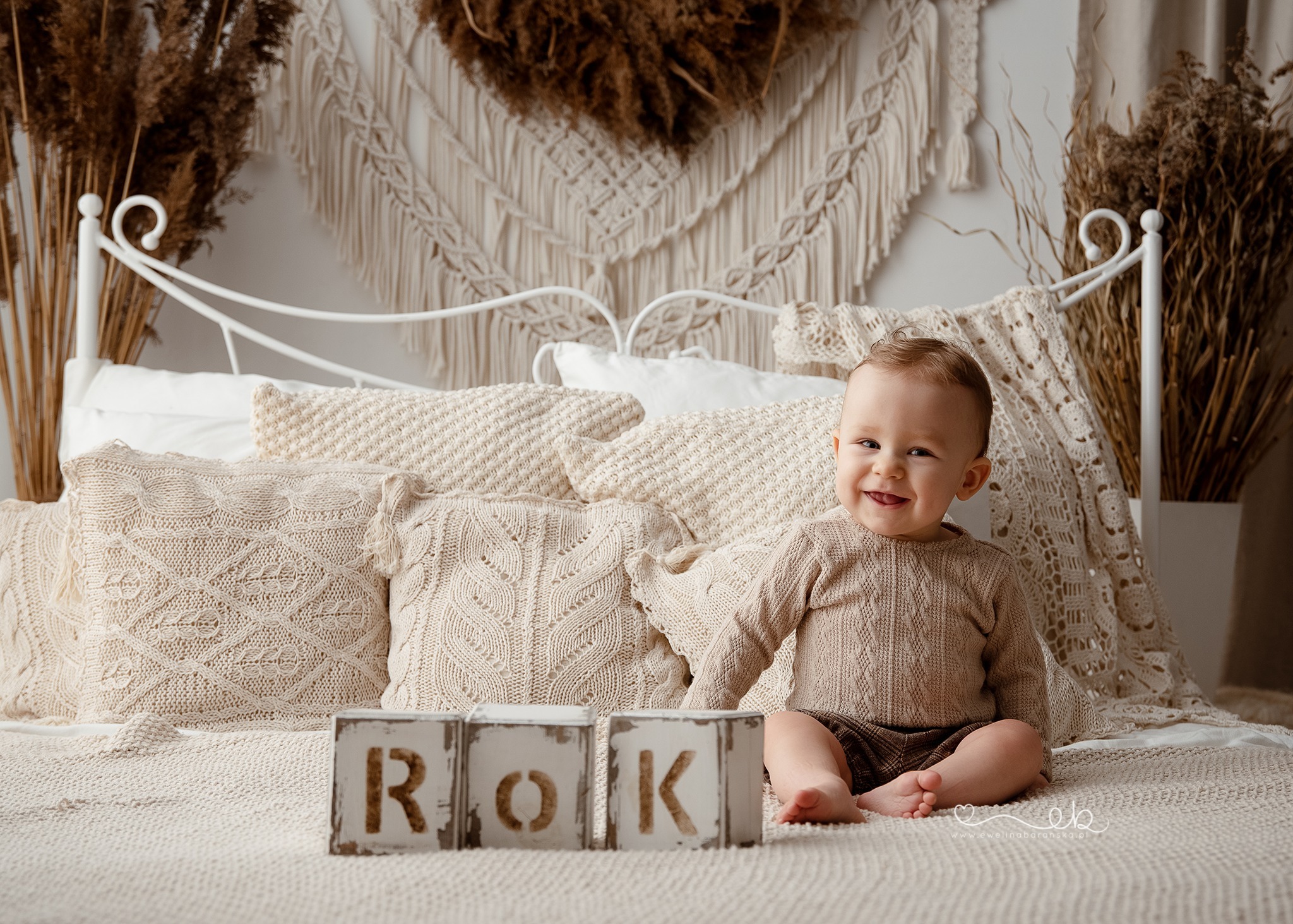 Uśmiechnięty roczny chłopiec siedzi na łóżku w beżowym swetrze i spodenkach, z drewnianymi klockami z napisem 'ROK' przed sobą, dekoracje z suszonych traw i makramy w tle.