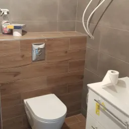 Nowoczesna łazienka z podwieszaną toaletą, drewnopodobnymi płytkami na podłodze i ścianie, oraz białą szafką pod umywalkę z widocznymi żółtymi etykietami, uzupełniona o grzejnik drabinkowy...