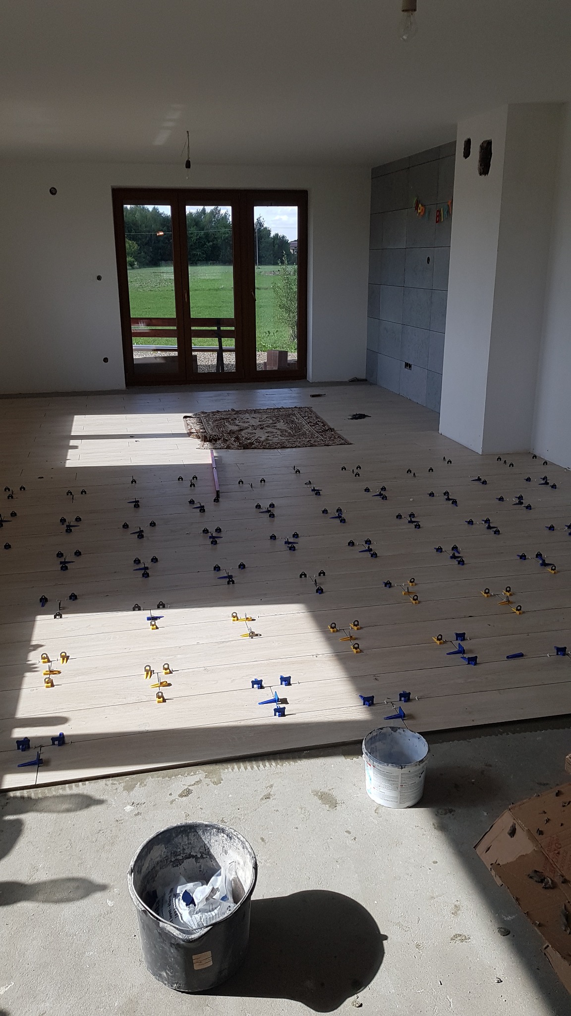 Jasne panele podłogowe w trakcie układania z użyciem klipsów poziomujących, widoczne dwa wiadra i wyjście na taras z widokiem na zieleń.