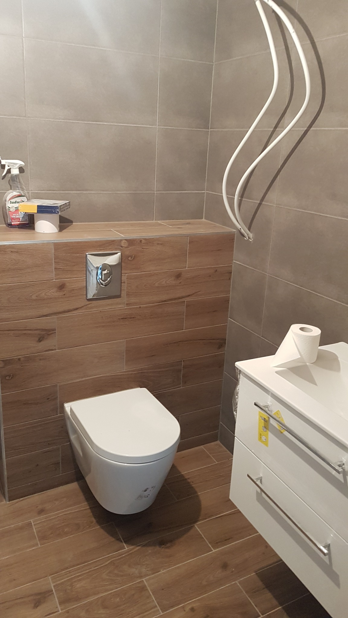 Nowoczesna łazienka z podwieszaną toaletą, drewnopodobnymi płytkami na podłodze i ścianie, oraz białą szafką pod umywalkę z widocznymi żółtymi etykietami, uzupełniona o grzejnik drabinkowy...