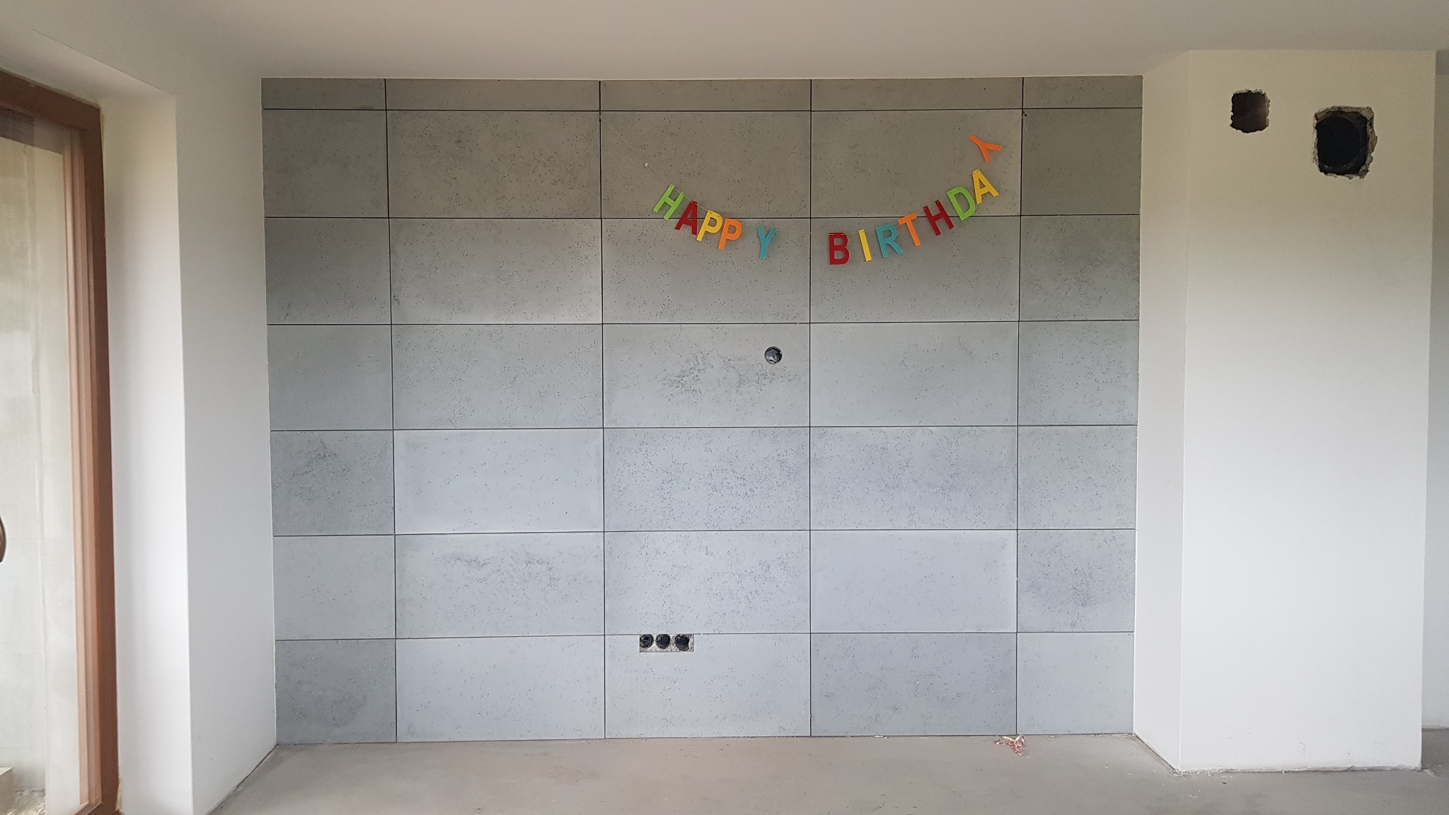 Ściana wyłożona szarymi płytami imitującymi beton, z kolorowym napisem 'Happy Birthday' zawieszonym na górze, widoczne puszki elektryczne w ścianie i fragment okna z drewnianą ramą.