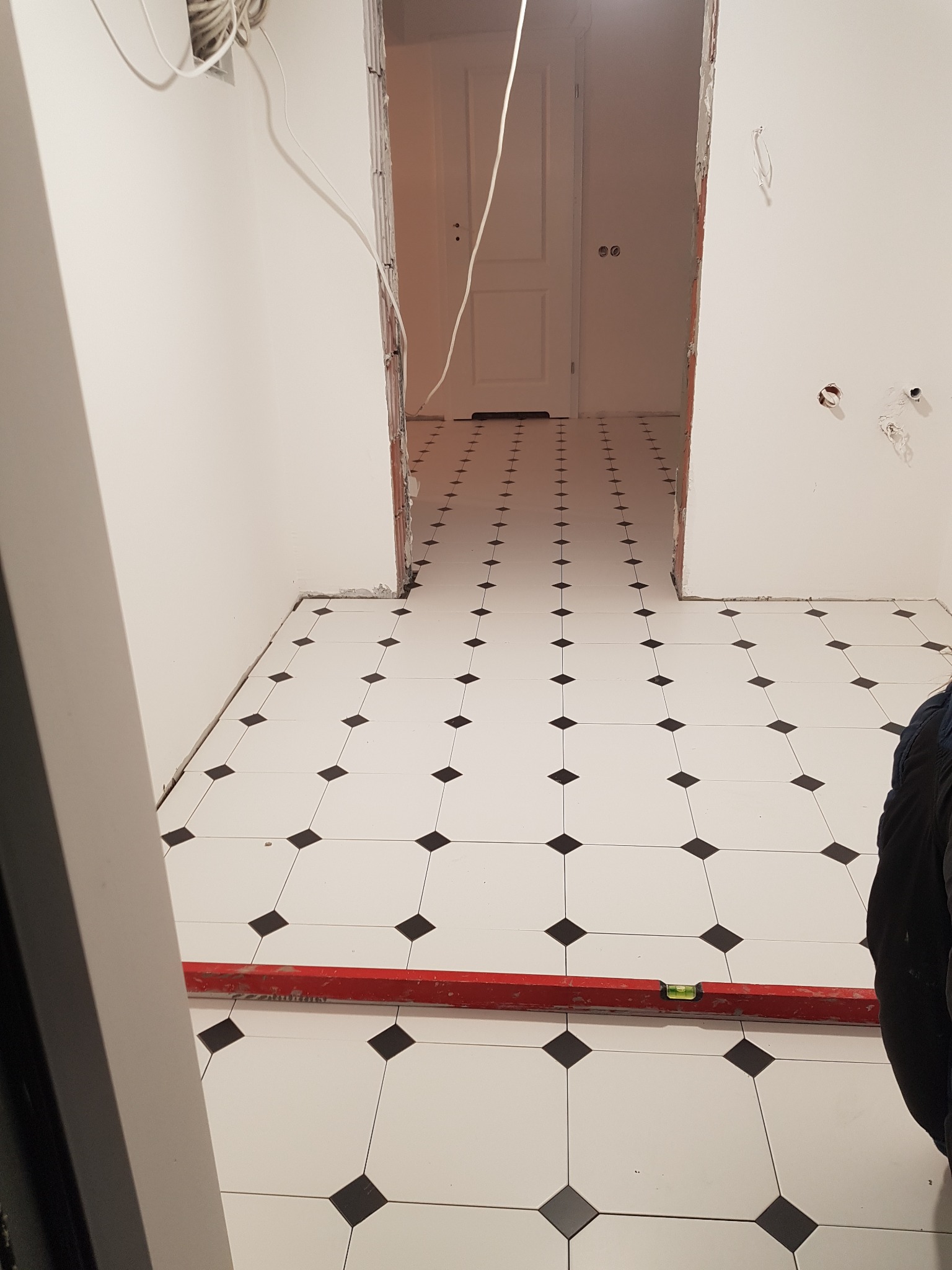 Ułożona podłoga z białych płytek z czarnymi rombami w nowym pomieszczeniu, widoczna poziomica, instalacja elektryczna w ścianie, brak listew przypodłogowych.