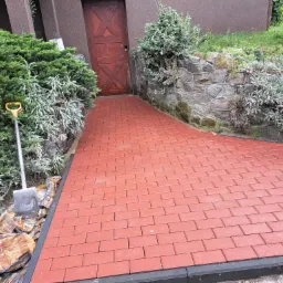 Czerwona kostka brukowa ułożona jako ścieżka do drewnianych drzwi, widoczna łopata z żółtym uchwytem i worki z cementem przy krawędzi.