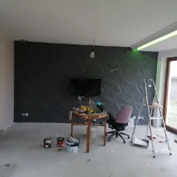 Pokój w trakcie remontu z ciemnoszarą ścianą z trójwymiarową fakturą, aluminiowa drabina oparta o ścianę, na podłodze puszki z farbą, wisząca żarówka bez klosza i okrągły stolik z narzędziami.