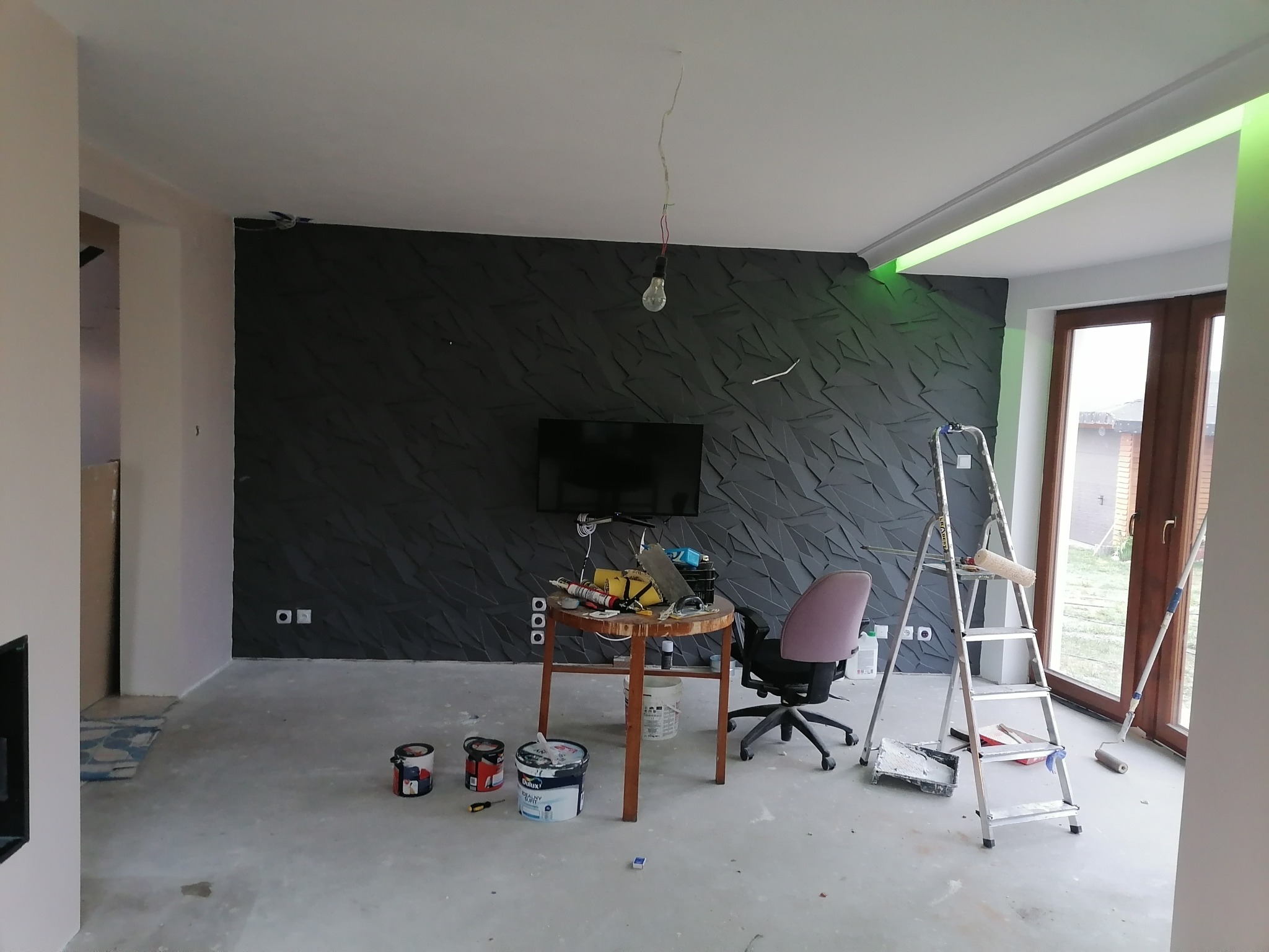 Pokój w trakcie remontu z ciemnoszarą ścianą z trójwymiarową fakturą, aluminiowa drabina oparta o ścianę, na podłodze puszki z farbą, wisząca żarówka bez klosza i okrągły stolik z narzędziami.