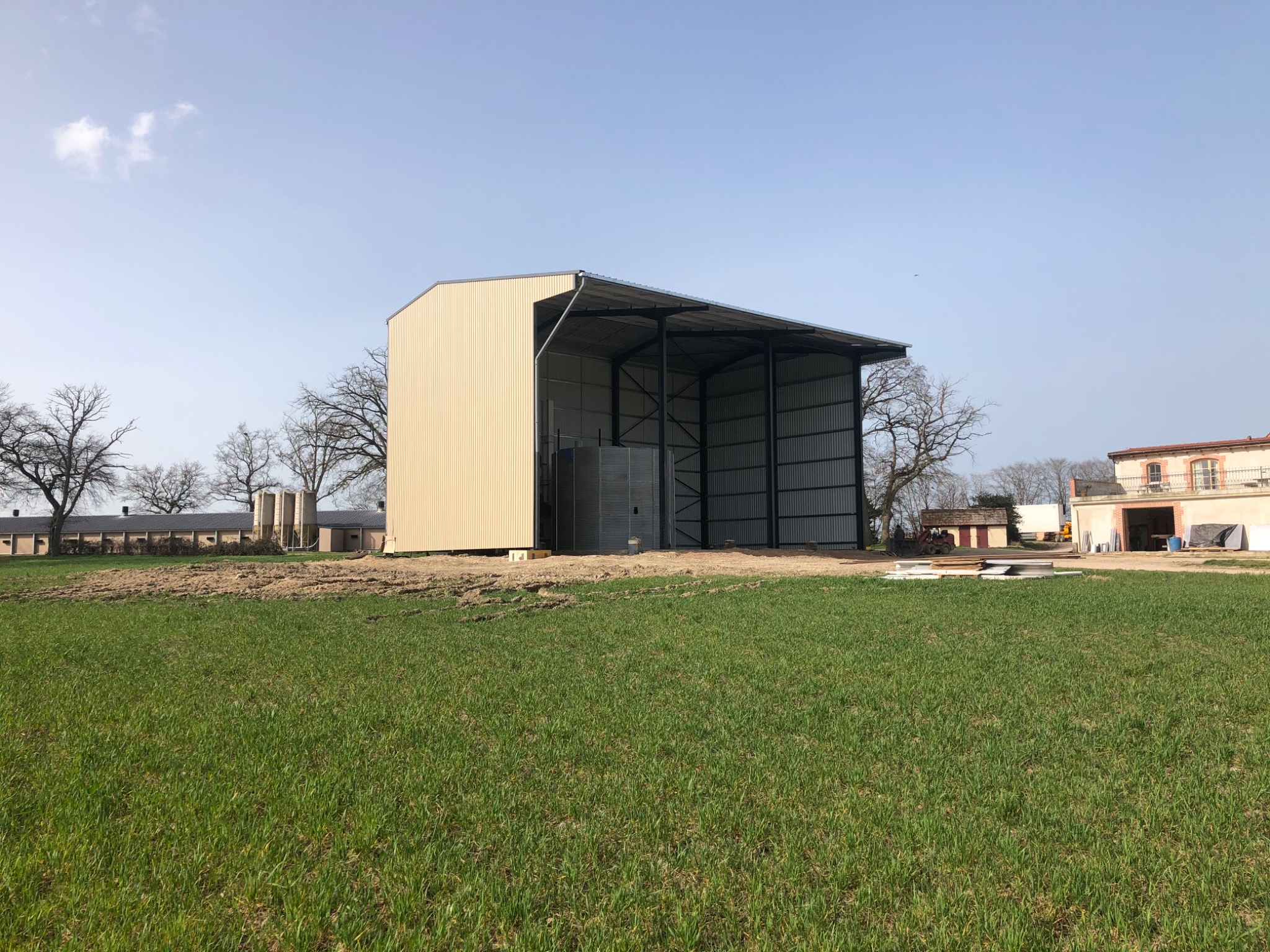 Szkielet konstrukcji stalowej z częściowo zamontowanymi płytami warstwowymi na tle zielonego pola i budynków gospodarczych.