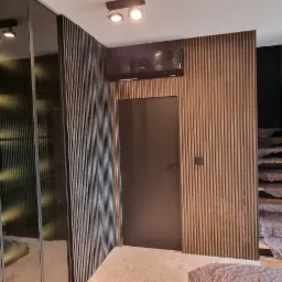 Wnętrze z szafą z lustrzanymi drzwiami, ścianą z drewnianych paneli, ciemnymi drzwiami i schodami zabezpieczonymi folią ochronną.