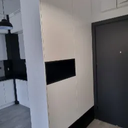 Nowoczesna biała szafa wnękowa z czarnymi akcentami, wkomponowana w przedpokój, widoczne drzwi wejściowe i fragment kuchni w tle.